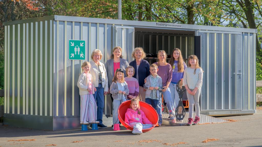 Gruppenbild der Lehrerinnen, Lehrer sowie Schülerinnen und Schüler vor ihrem neuen Spielzeug- und Materialcontainer auf dem Schulhof der Grundschule Dattenfeld.