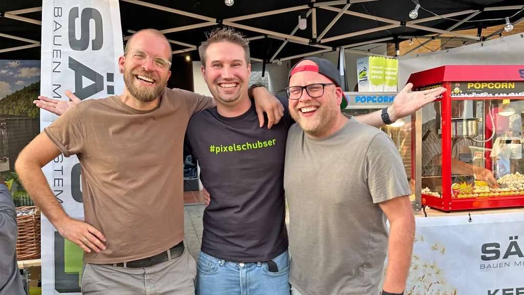 Drei herzhafte lachende Männer, einer im "#pixelschubser"-T-Shirt, stehen Arm in Arm vor einem Pavillon mit einem Popcornautomaten.